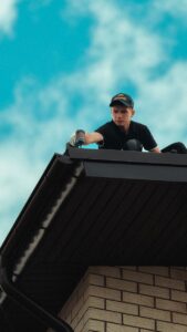 Roofer working diligently on a house roof under a bright blue sky. Professional roofing maintenance in progress.